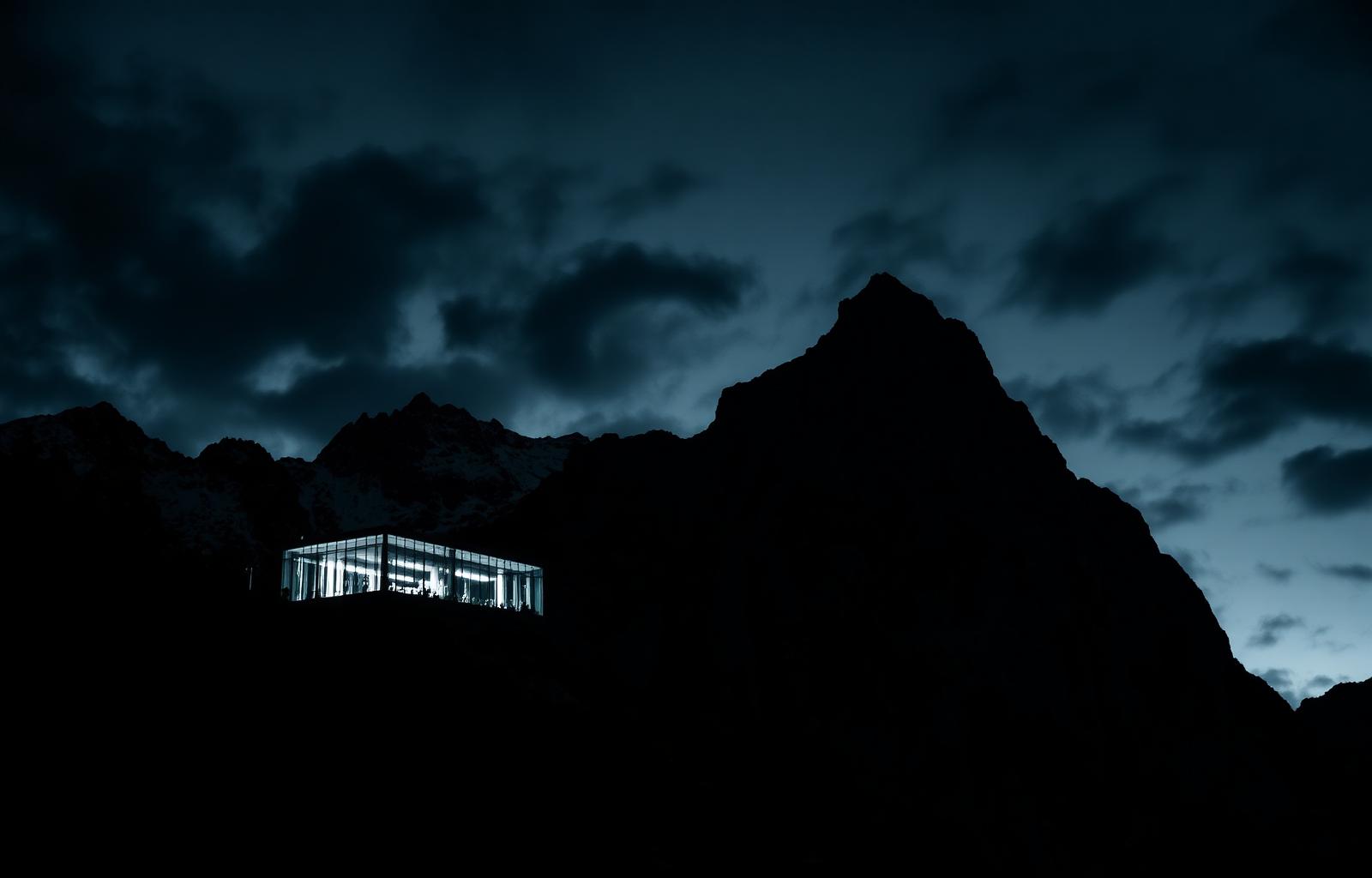 A modern glass research facility set into the Swiss Alps at dusk, photographed in dramatic black and white.